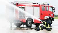 Feuerwehrmänner der Bundeswehr knien mit einem Wasserschlauch in der Hand, im Hintergrund ein Feuerwehrfahrzeug Gebäudebrand.