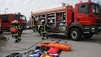 Rettungsmittel und Schläuche der Feuerwehr sind auf dem Boden ausgebreitet, Feuerwehrmänner bewegen sich vor zwei Fahrzeugen.