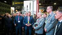 Männer in Anzügen und Uniformen stehen auf einer Messe zwischen Ständen. Ein Zivlist spricht.