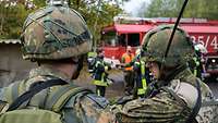 Im Hintergrund steht ein Löschfahrzeug mit Brandschützern, im Vordergrund zwei Sicherungssoldaten.