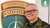 Der Militärseelsorger steht vor einem Gebäude. Hinter ihm an der Wand hängt das Wappen der Enhanced Forward Presence