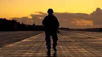 Ein Objektschützer geht mit seinem Gewehr im Sonnenuntergang die Landebahn entlang und sichert sie.