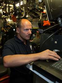 In einem halbdunklen Raum: Ein Marinesoldat in blauem Shirt arbeitet stehend an einem Laptop; im Hintergrund Rohre und Monitore.