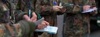 Mehrere Soldaten stehen mit Schreibblöcken und Stiften im Halbkreis und schreiben.