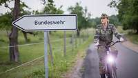 Eine Soldatin fährt auf einem Fahrrad an einem Schild mit der Aufschrift „Bundeswehr“ vorbei.