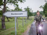 Eine Soldatin fährt auf einem Fahrrad an einem Schild mit der Aufschrift „Bundeswehr“ vorbei.