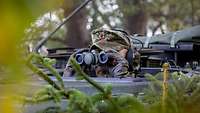 Ein Soldat schaut an einem Waldrand mit einem Fernglas aus einer Panzerluke ins Gelände.