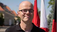 Manfred Freuding im Portraitvor einer polnischen, dänischen und deutschen Flagge. Im Hintergrund ein Kasernengebäude.