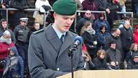 Ein Soldat, hinter einem Pult stehend, hält eine Rede. Auf der Tribüne dahinter stehen Gäste.