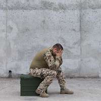 Ein Soldat sitzt auf einer Metallkiste und hält die Hände vor das Gesicht und stützt die Ellenbogen auf den Beinen ab