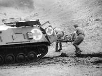 Soldaten des Sanitätsdienstes bei einer Übung mit dem Panzer Hotchkiss Schwarzweiß-Aufnahme: Zwei Soldaten laufen mit einem Verletzten auf einer Trage zum Sanitätspanzer Hotchkiss.