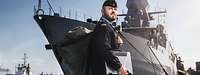 Ein Marinesoldat im blauen Arbeitsanzug im Hafen, im Hintergrund ein graues Kriegsschiff an der Pier.