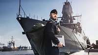 Ein Marinesoldat im blauen Arbeitsanzug im Hafen, im Hintergrund ein graues Kriegsschiff an der Pier.