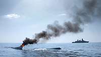 Fregatte Hamburg Operation Atalanta Auf einer Wasseroberfläche brennt ein fast versunkenes Boot, im Hintergrund ein Kriegsschiff.