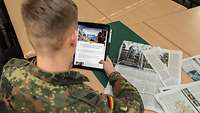 Ein Soldat sitzt an einem Tisch mit Zeitschriften und hält ein Tablet in der Hand.