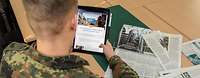Ein Soldat sitzt an einem Tisch mit Zeitschriften und hält ein Tablet in der Hand.