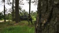 Ein Soldat läuft zwischen Bäumen im Wald, im Hintergrund ein Schützenpanzer.