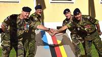 Vier Soldaten legen jeweils eine Hand zur Mitte hin übereinander, im Hintergrund das Brigadewappen..