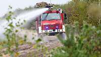 Ein Waldbrandlöschfahrzeug fährt auf einem Feldweg, Düsen befeuchten den Weg, ein Feuerwehrmann am Dachwerfer. 