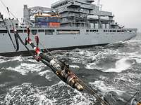 Einsatzgruppenversorger Bonn RAS Ein großes graues Schiff in See, von einem Ladebaum aus hängen eine Stahltrosse und ein schwarzer Schlauch nach vorne ins Bild.