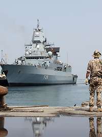 Marine im Einsatz Ein graues Kriegsschiff hält auf eine Kaimauer zu; auf der Kaimauer zwei Soldaten in beigefarbener Flecktarnuniform.