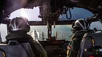 Blick aus einem Hubschrauber-Cockpit, mit Pilot und Co-Pilot; im Fenster ein Kriegsschiff auf See fast direkt von oben