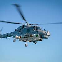Ein dunkelgrauer Hubschrauber im Flug von halb unten; seitlich unter dem Rumpf zwei kurze Torpedos