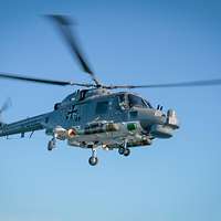 Ein dunkelgrauer Hubschrauber im Flug von halb unten; seitlich unter dem Rumpf zwei kurze Torpedos