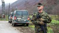 Ein Soldat steht am Straßenrand. Hinter ihm stehen zwei grüne Transporter mitde Aufschrift KFOR.