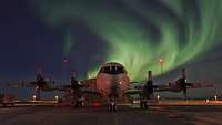 Ein stehendes, viermotoriges Propellerflugzeug. Es ist dunkel, im Hintergrund an Himmel grüne Lichtschleier des Nordlichts.