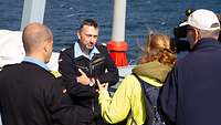 Ein Admiral in blauer Arbeitsuniform, vor ihm eine Frau mit grüngelber Windjacke und ein Mann mit einer Videokamera.