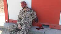 Ein Soldat mit abgerissenem Arm sitzt an einer Containerwand 