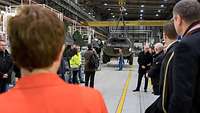 Eine Frau in rotem Blazer schaut in eine Werkhalle. Dort hängt an Stahlseilen ein Militärfahrzeug, 