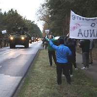 Auf einer Landstraße fahren Militärfahrzeuge, am Straßenrand stehen Zivilisten mit Transparenten.