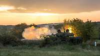 Ein Panzer steht in einer getarnten Stellung und schießt. Vor ihm scheinen Feuerwolken auf.