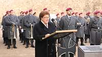 Eine Frau im schwarzen Mantel spricht an einem Rednerpult, hinter ihr Soldaten mit roten Baretten.