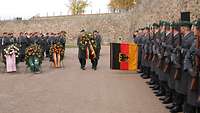 Soldaten mit Waffen sind an einer Festungsmauer angetreten. Zwei Soldaten tragen einen Trauerkranz.