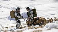 Zwei Soldaten lassen im Schnee eine spezielle Trage mit Kufen den Hang herunter.