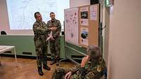 Ein Soldat deutet in einem Raum mit einem Zeigestock auf Fotos und eine Karte an einer Pinnwand.