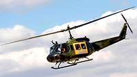 Ein orange-grüner Hubschrauber fliegt unter leicht bewölktem Himmel.