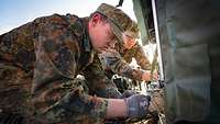 Zwei Soldaten hocken am Boden und ziehen Kabel an einer Zeltwand entlang.