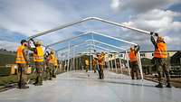 Neun Soldaten stehen auf Holzböden und bauen Zelte aus Aluminiumgestellen auf.