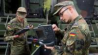 Zwei Soldaten halten Telefone in der Hand. Im Hintergrund stehen zwei Lkw mit Kabinen.