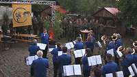 Ein Orchester gekleidet in blauen Anzügen spielt stehend im Freien, zahlreiche Soldaten hören zu.