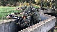 Soldaten stehen am Waldrand in einem Betongraben und haben ihre Gewehre auf Sandsäcke gelegt.