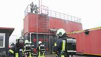 Personen in Schutzanzügen mit Schutzhelmen und Atemluftlflaschen stehen vor roten und schwarzen Containern.