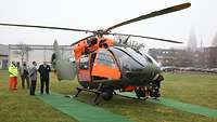 Ein Hubschrauber steht auf einer Rasenfläche, im Hintergrund vier Männer.
