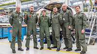 Sechs Soldaten in Fliegeruniform stehen in der Werkshalle vor einem Rumpf des neuen Hubschraubers.