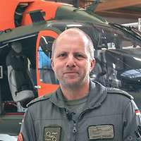 Ein Hubschrauberpilot steht lächelnd vor einem Rettungshubschrauber mit orangefarbenen Aufklebern.