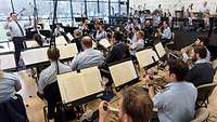 Ein Dirigent steht vor Musikern in einem großen Raum.